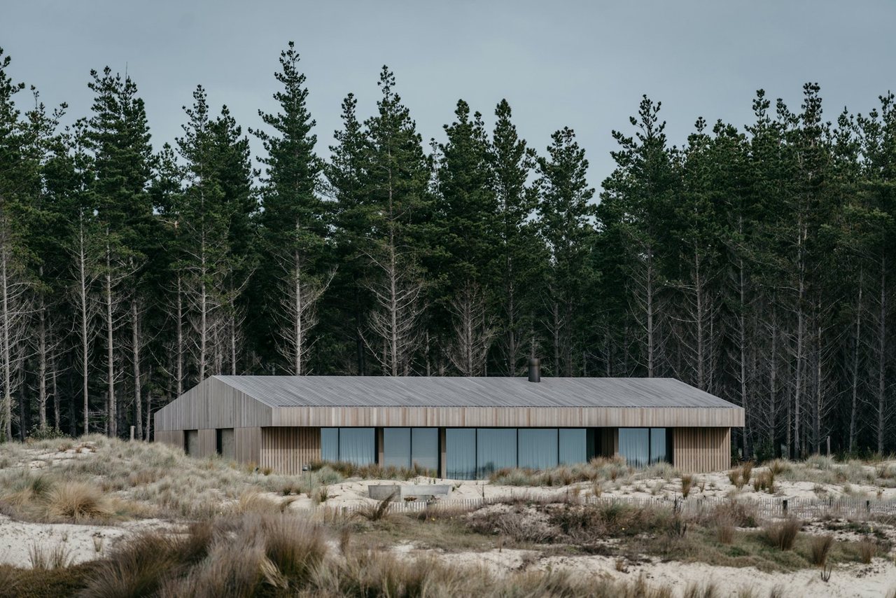 A low single-storey timber house with a shallow metal roof standing among coastal sand dunes and marram grass, backed by a dense stand of pine trees under an overcast New Zealand sky