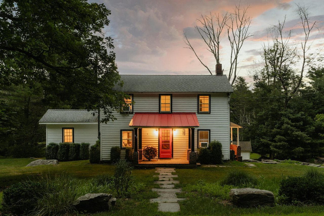 A modest two-storey white clapboard house at dusk, porch light on under a small red awning, set back from a stone path across a lawn with woodland behind, the kind of older starter home that sits below the median US price