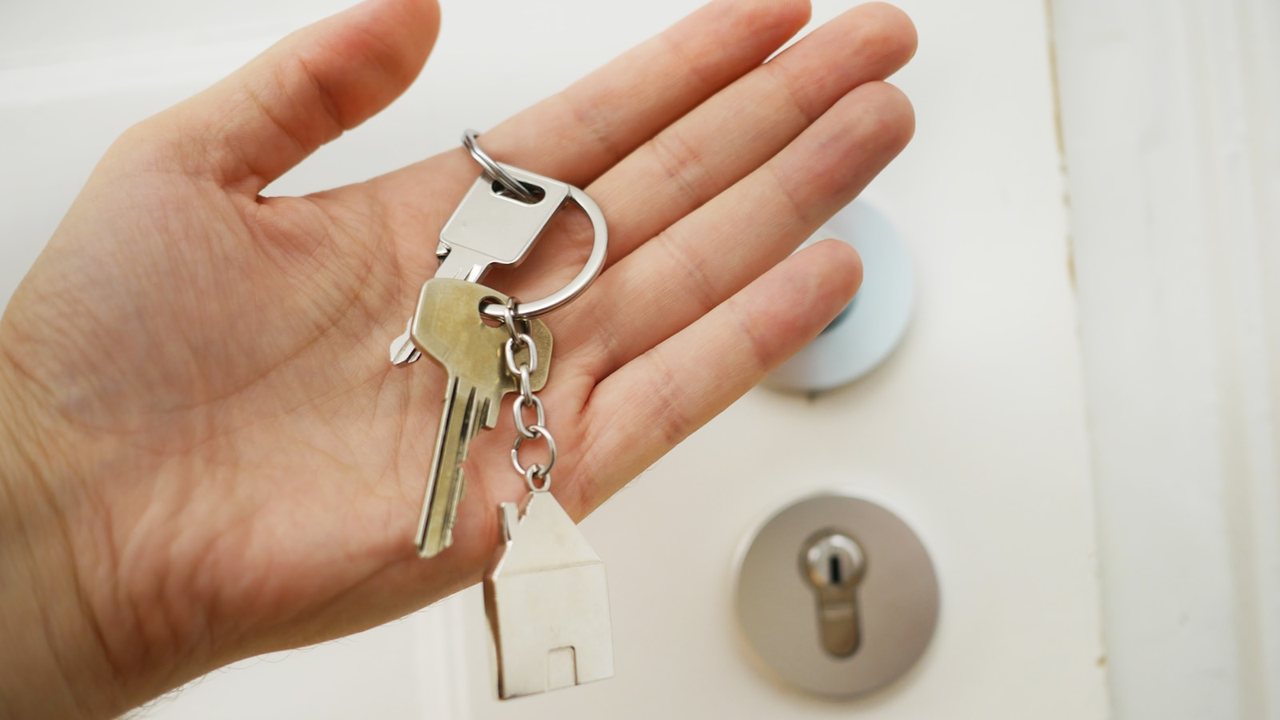 An open hand holding a set of house keys with a small house-shaped keyring, in front of a white front door with a lock